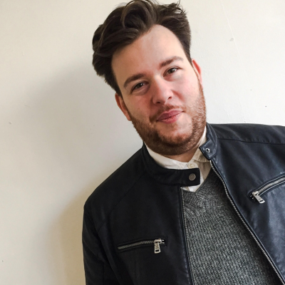 A white man with dark hair wearing a grey jumper and a leather jacket smiles at the camera.